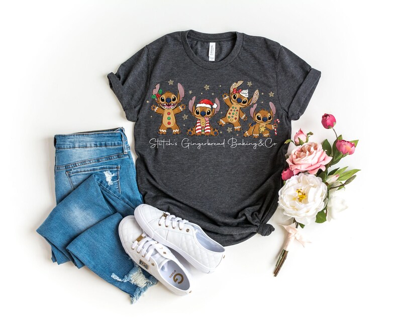 Puede incluir: Camiseta gris oscuro con cuatro personajes de dibujos animados de pan de jengibre, adornados con accesorios festivos. El texto "Stitch's Gingerbread Baking & Co" est&aacute; impreso debajo. La camiseta se combina con vaqueros azules y zapatillas blancas.