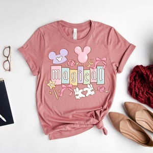 May include: A dusty rose t-shirt with the word "magical" in colorful block letters. The design includes Mickey Mouse ears, a magic wand, bows, roses, and a heart-shaped bunny. The shirt is folded on a white surface.