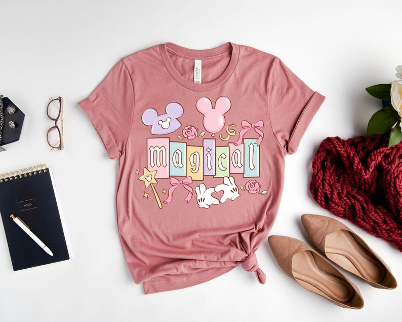 May include: A dusty rose t-shirt with the word "magical" in colorful block letters. The design includes Mickey Mouse ears, a magic wand, bows, roses, and a heart-shaped bunny. The shirt is folded on a white surface.