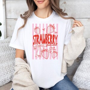May include: White t-shirt featuring a red and pink strawberry design. The graphic includes the words "STRAWBERRY FESTIVAL" and strawberry illustrations, along with the phrase "a berry good time". The shirt has a crew neck and short sleeves.