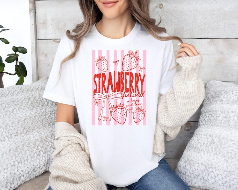May include: White t-shirt featuring a red and pink strawberry design. The graphic includes the words "STRAWBERRY FESTIVAL" and strawberry illustrations, along with the phrase "a berry good time". The shirt has a crew neck and short sleeves.