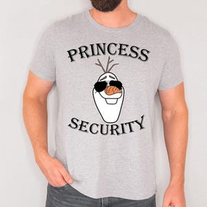 Pu&ograve; includere: T-shirt grigio chiaro con le parole "PRINCESS SECURITY" sopra e sotto un pupazzo di neve dei cartoni animati con occhiali da sole e un naso a carota. La maglietta &egrave; a maniche corte.