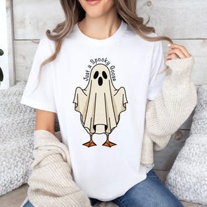 May include: White t-shirt featuring a cartoon ghost with goose feet and the text "Just a Spooky Goose" in a circular font. The ghost is beige with black eyes and a mouth. The goose feet are orange.