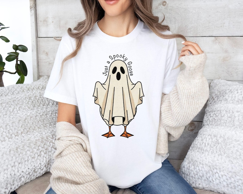 May include: White t-shirt featuring a cartoon ghost with goose feet and the text "Just a Spooky Goose" in a circular font. The ghost is beige with black eyes and a mouth. The goose feet are orange.