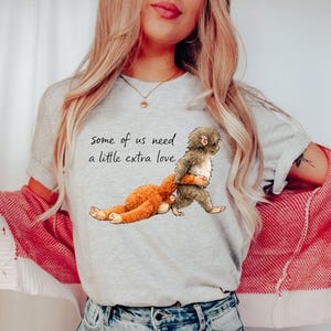 Peut inclure: T-shirt gris clair avec un dessin de deux singes de dessin anim&eacute; qui s'embrassent. Le texte "some of us need a little extra love" est imprim&eacute; au-dessus des singes. Le t-shirt est fait d'une mati&egrave;re douce.