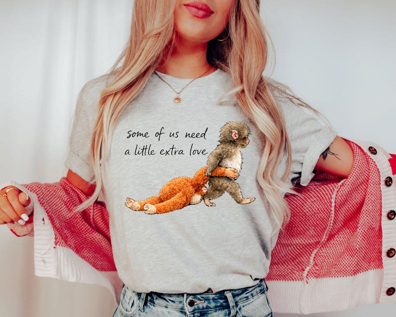 Peut inclure: T-shirt gris clair avec un dessin de deux singes de dessin anim&eacute; qui s'embrassent. Le texte "some of us need a little extra love" est imprim&eacute; au-dessus des singes. Le t-shirt est fait d'une mati&egrave;re douce.