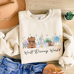 May include: Cream-colored sweatshirt featuring a colorful illustration of Walt Disney World landmarks, including Cinderella's castle and the Tower of Terror. The words "Walt Disney World" are written in a script font below the graphic. The sweatshirt is paired with blue denim shorts.