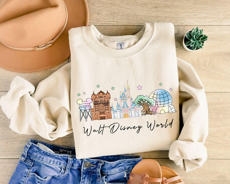 May include: Cream-colored sweatshirt featuring a colorful illustration of Walt Disney World landmarks, including Cinderella's castle and the Tower of Terror. The words "Walt Disney World" are written in a script font below the graphic. The sweatshirt is paired with blue denim shorts.