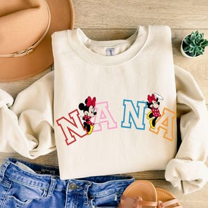 May include: A cream-colored sweatshirt with the word "NANA" in rainbow letters and two cartoon Mickey Mouse characters on either side of the word.
