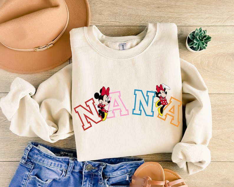 May include: A cream-colored sweatshirt with the word "NANA" in rainbow letters and two cartoon Mickey Mouse characters on either side of the word.