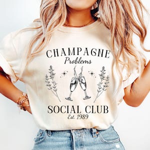 May include: A white t-shirt with the text "Champagne Problems Social Club Est. 1989" and a graphic of two champagne glasses with a floral design.