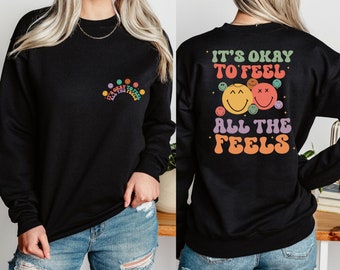 Puede incluir: Sudadera negra con un diseño de cara sonriente arcoíris y el texto "It's okay to feel all the feels".
