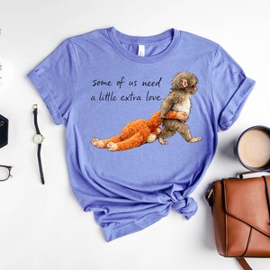 Peut inclure: Un t-shirt bleu clair avec le texte "some of us need a little extra love." Le t-shirt pr&eacute;sente un dessin d'un singe portant un animal en peluche. Autres accessoires : une montre, des lunettes et un sac &agrave; main.