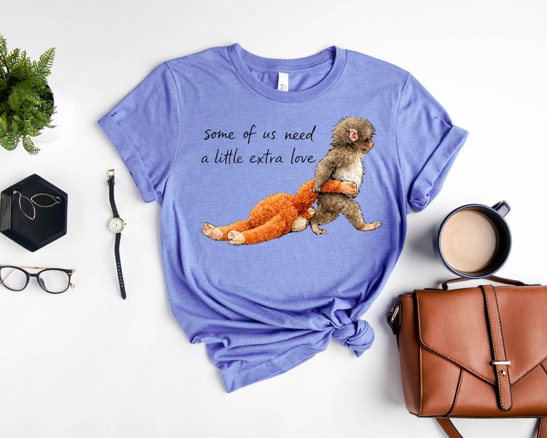 Peut inclure: Un t-shirt bleu clair avec le texte "some of us need a little extra love." Le t-shirt pr&eacute;sente un dessin d'un singe portant un animal en peluche. Autres accessoires : une montre, des lunettes et un sac &agrave; main.