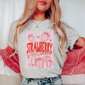 May include: A light grey t-shirt featuring a red and pink graphic with the words "Strawberry Festival" and strawberry illustrations. The shirt has a crew neck and short sleeves. The text "a berry good time" is also included.