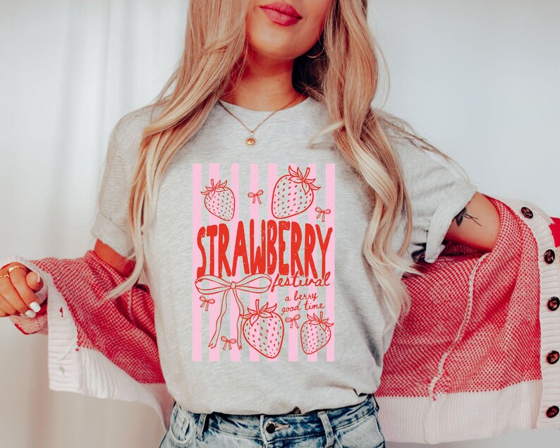 May include: A light grey t-shirt featuring a red and pink graphic with the words "Strawberry Festival" and strawberry illustrations. The shirt has a crew neck and short sleeves. The text "a berry good time" is also included.