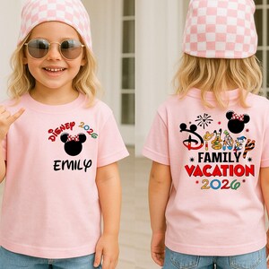 May include: A light pink t-shirt with the text "Disney 2026" and a Minnie Mouse graphic. The front of the shirt has the name "Emily" printed below the graphic. The back of the shirt says "Family Vacation 2026".