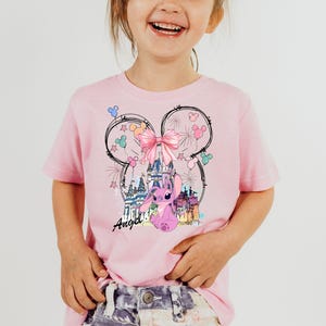 May include: Pink t-shirt featuring a graphic of a cartoon character with a castle, bow, and fireworks design. The word "Angel" is written in cursive. The shirt is a light pink color.