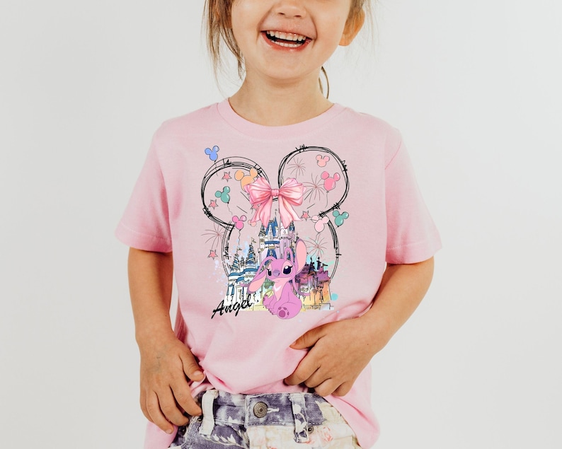 May include: Pink t-shirt featuring a graphic of a cartoon character with a castle, bow, and fireworks design. The word "Angel" is written in cursive. The shirt is a light pink color.
