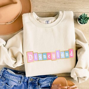 Puede incluir: Sudadera color crema con la palabra "Disneyland" en colores pastel y bordes dorados. La sudadera se combina con vaqueros azules, un sombrero beige y sandalias. Tambi&eacute;n se ve una peque&ntilde;a planta suculenta en maceta.