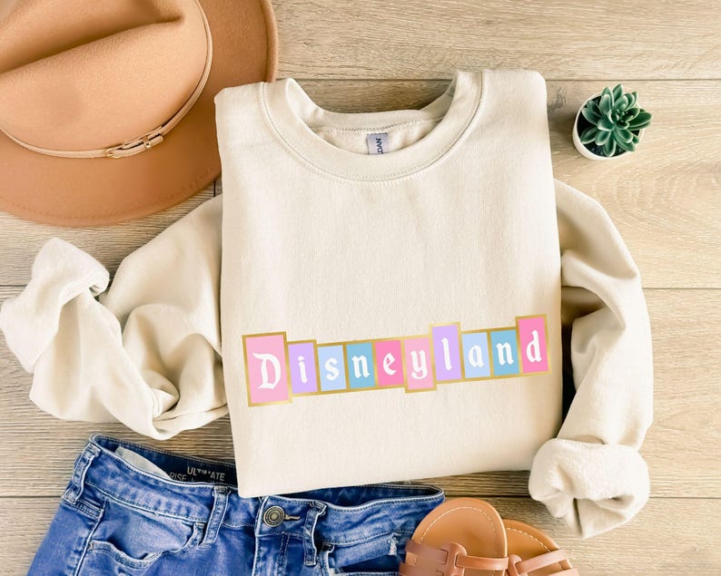 Puede incluir: Sudadera color crema con la palabra "Disneyland" en colores pastel y bordes dorados. La sudadera se combina con vaqueros azules, un sombrero beige y sandalias. Tambi&eacute;n se ve una peque&ntilde;a planta suculenta en maceta.