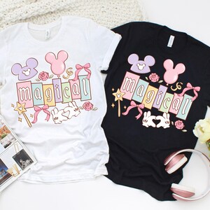 May include: Two t-shirts, one white and one black, each featuring a colorful graphic. The graphic includes the word "magical" in pastel colors, Mickey Mouse-inspired shapes, a wand, bows, roses, and a pair of bunny hands forming a heart.