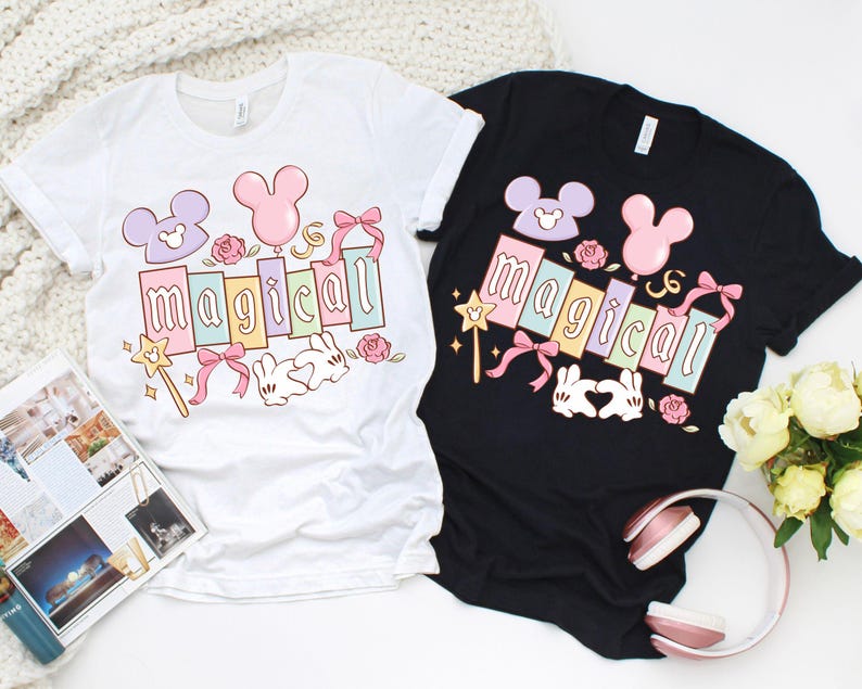 May include: Two t-shirts, one white and one black, each featuring a colorful graphic. The graphic includes the word "magical" in pastel colors, Mickey Mouse-inspired shapes, a wand, bows, roses, and a pair of bunny hands forming a heart.