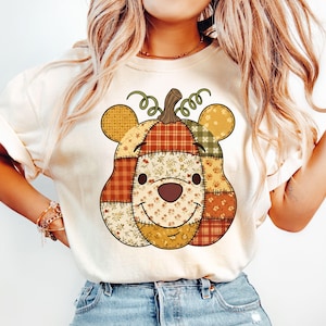以下が含まれることがあります： クリーム色のTシャツ。パッチワークのパンプキンデザイン、クマの耳、笑顔の顔が特徴です。カボチャはオレンジ、黄色、緑の様々なパターンで構成され、茶色の茎とカールした巻きひげが付いています。カジュアルで秋に適したシャツです。