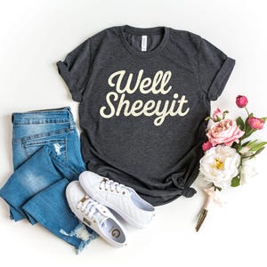 May include: A dark grey t-shirt with the text "Well Sheeeyit" in a retro font. The image includes blue jeans, white trainers, and a bouquet of pink and white flowers, arranged on a white surface.