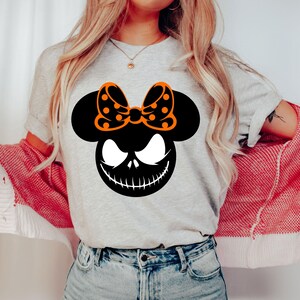 Puede incluir: Camiseta gris claro con una cabeza de Mickey Mouse negra con una cara de Jack Skellington y un lazo naranja con lunares. Dise&ntilde;o tem&aacute;tico de Halloween.