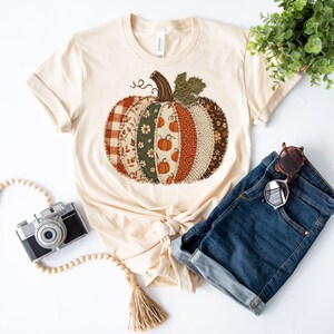 May include: A cream-coloured short-sleeve t-shirt featuring a large central graphic of a pumpkin composed of various fabric-like patterns including plaid, floral, and herringbone prints in shades of orange, green, brown, and white.