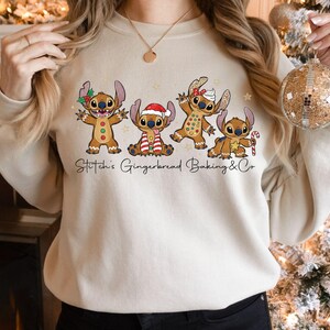 Puede incluir: Sudadera beige con un gr&aacute;fico de cuatro personajes Stitch estilo pan de jengibre con atuendos festivos. El texto "Stitch's Gingerbread Baking & Co" est&aacute; impreso debajo. La sudadera es de color neutro.