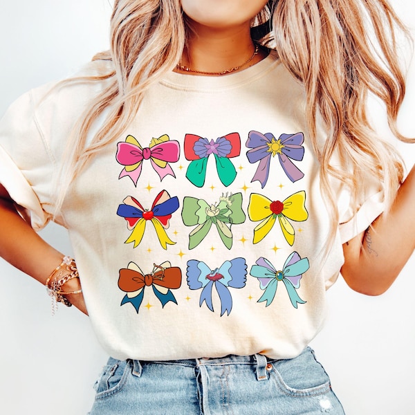 以下が含まれることがあります： クリーム色のTシャツ。カラフルな漫画のリボンが格子状に並んでいます。リボンはピンク、赤、緑、紫、黄色、青など様々な色があります。遊び心があり、気まぐれなデザインです。