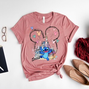 May include: A dusty rose t-shirt featuring a graphic of Stitch from Lilo & Stitch in front of a castle, with Mickey Mouse ears and fireworks. The word "Stitch" is written below the character. The shirt is styled with accessories.