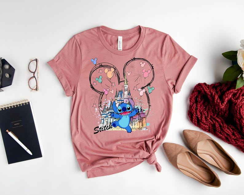 May include: A dusty rose t-shirt featuring a graphic of Stitch from Lilo & Stitch in front of a castle, with Mickey Mouse ears and fireworks. The word "Stitch" is written below the character. The shirt is styled with accessories.