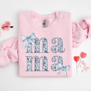 K&ouml;nnte beinhalten: Rosafarbener Pullover mit einem blauen und wei&szlig;en Blumenmuster, das "mama" buchstabiert, mit einer blauen Schleife unten.