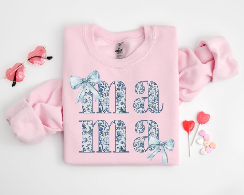K&ouml;nnte beinhalten: Rosafarbener Pullover mit einem blauen und wei&szlig;en Blumenmuster, das "mama" buchstabiert, mit einer blauen Schleife unten.