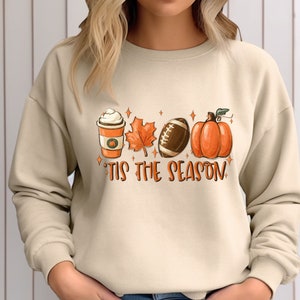 K&ouml;nnte beinhalten: Ein beiger Pullover mit einem grafischen Aufdruck einer Kaffeetasse, einem Blatt, einem Football und einem K&uuml;rbis. Der Text "Tis the Season" ist unter dem Aufdruck gedruckt.