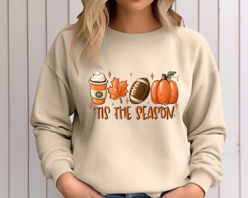 K&ouml;nnte beinhalten: Ein beiger Pullover mit einem grafischen Aufdruck einer Kaffeetasse, einem Blatt, einem Football und einem K&uuml;rbis. Der Text "Tis the Season" ist unter dem Aufdruck gedruckt.
