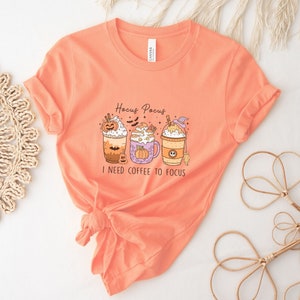 May include: Orange t-shirt with a graphic of three Halloween-themed coffee drinks with the text "Hocus Pocus I need coffee to focus".
