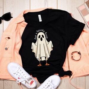 May include: Black t-shirt with a ghost graphic and the text "Just a Spooky Goose." The shirt is paired with a peach-colored jacket, white sneakers, and black sunglasses. A Halloween-themed design.