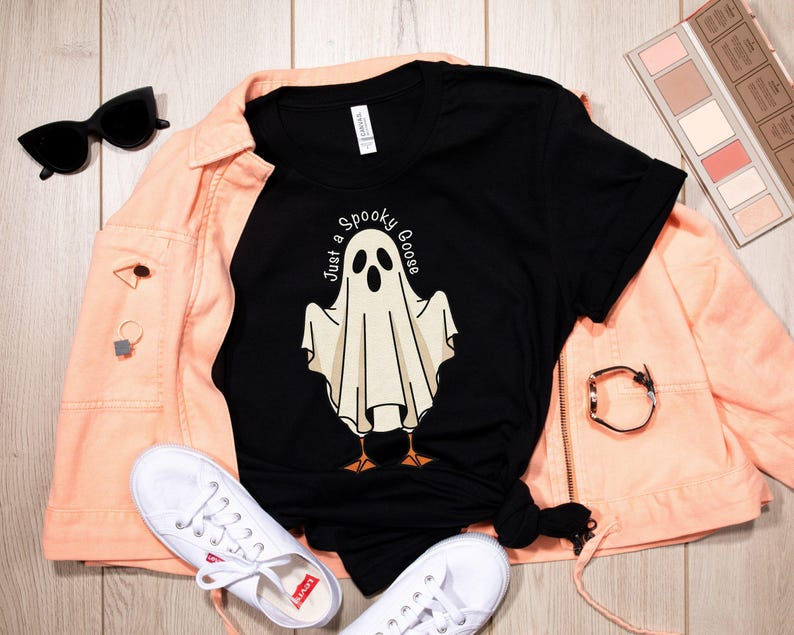 May include: Black t-shirt with a ghost graphic and the text "Just a Spooky Goose." The shirt is paired with a peach-colored jacket, white sneakers, and black sunglasses. A Halloween-themed design.