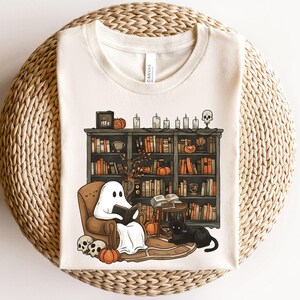 Könnte beinhalten: Cremefarbenes T-Shirt mit einer Halloween-Illustration. Ein Geist liest in einem Sessel vor einem Bücherregal mit Büchern, Kürbissen und Kerzen. Eine schwarze Katze sitzt in der Nähe. Schädel und Kürbisse liegen auf dem Boden.