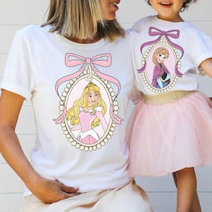 以下が含まれることがあります： ピンクのリボンとフレームの中にプリンセスが描かれた白いTシャツ2枚。左側のプリンセスは眠れる森の美女のオーロラ。右側のプリンセスはアナと雪の女王のエルサ。Tシャツは母娘用です。