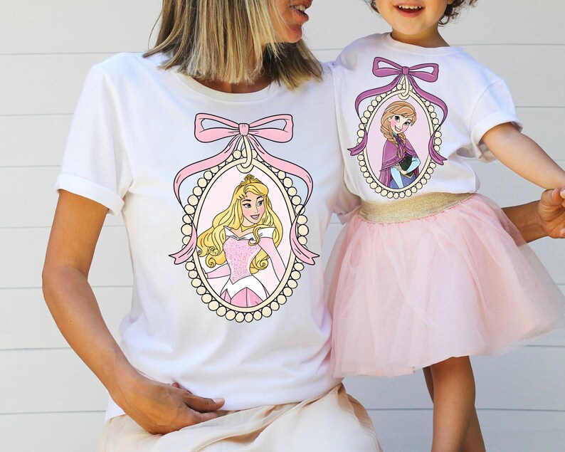 以下が含まれることがあります： ピンクのリボンとフレームの中にプリンセスが描かれた白いTシャツ2枚。左側のプリンセスは眠れる森の美女のオーロラ。右側のプリンセスはアナと雪の女王のエルサ。Tシャツは母娘用です。