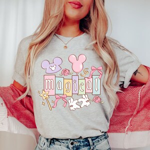 May include: A light gray t-shirt with the word "magical" in pastel colors, decorated with cartoon elements like a wand, bows, and a Mickey Mouse head. The shirt is worn by a person with long blonde hair.