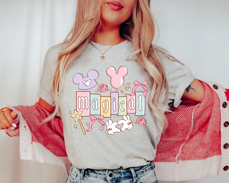 May include: A light gray t-shirt with the word "magical" in pastel colors, decorated with cartoon elements like a wand, bows, and a Mickey Mouse head. The shirt is worn by a person with long blonde hair.