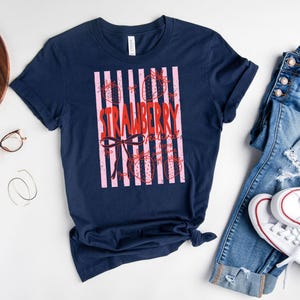 May include: A navy blue t-shirt with a pink and red strawberry design. The word "STRAWBERRY" is written in red, with a bow and strawberry illustrations. A casual tee for everyday wear.