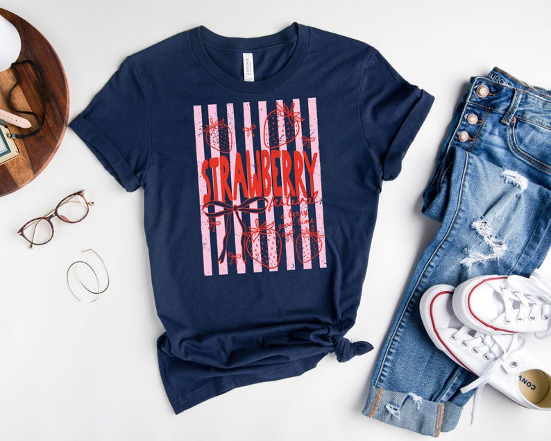 May include: A navy blue t-shirt with a pink and red strawberry design. The word "STRAWBERRY" is written in red, with a bow and strawberry illustrations. A casual tee for everyday wear.