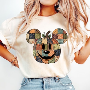 May include: Cream-colored t-shirt featuring a patchwork pumpkin design with Mickey Mouse ears. The pumpkin is composed of various fabric patterns in orange, green, and brown. The design has a smiling face and a stem with leaves.